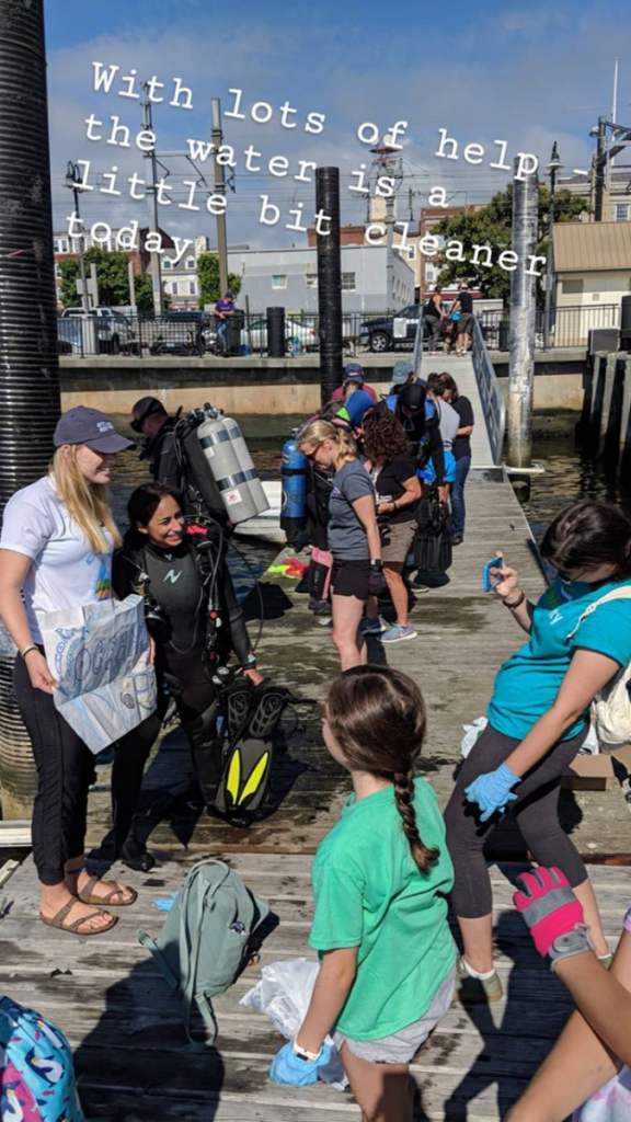 SCUBA divers and kids pose together on a pier; overlay text says "with lots of  help, the water is a little bit cleaner today"