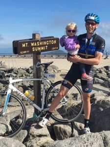 David Anderson posing with a toddler and bike and a Mt Washington summit sign