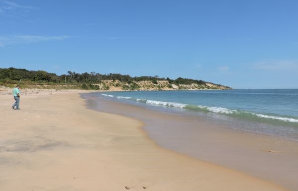 photo of a wide beach with bluffs at the back. It's a sunny day with blue sky and aqua water. There is a single person in jeans walking on the beach in the distance.