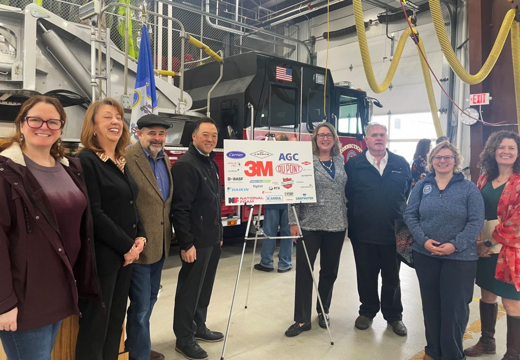 Several advocates stand around a sign with the logos of companies being sued for PFAS pollution